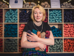 A blond haired woman holding an armful of coloured wool balls standing in front of a display of the same wool.